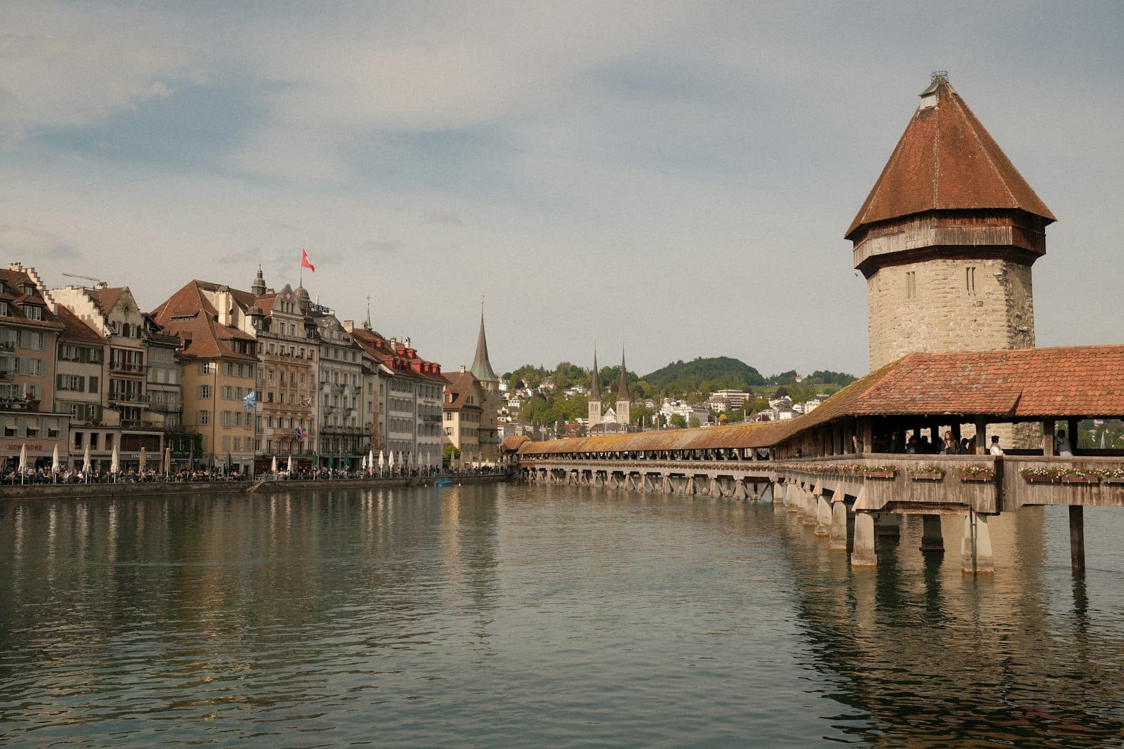 วิวกว้างของสะพาน Chapel Bridge และหอคอย Water Tower แห่ง Lucerne ที่ทอดข้ามแม่น้ำ พร้อมอาคารประวัติศาสตร์ในย่านเมืองเก่าริมน้ำใต้ท้องฟ้ามีเมฆบางส่วน