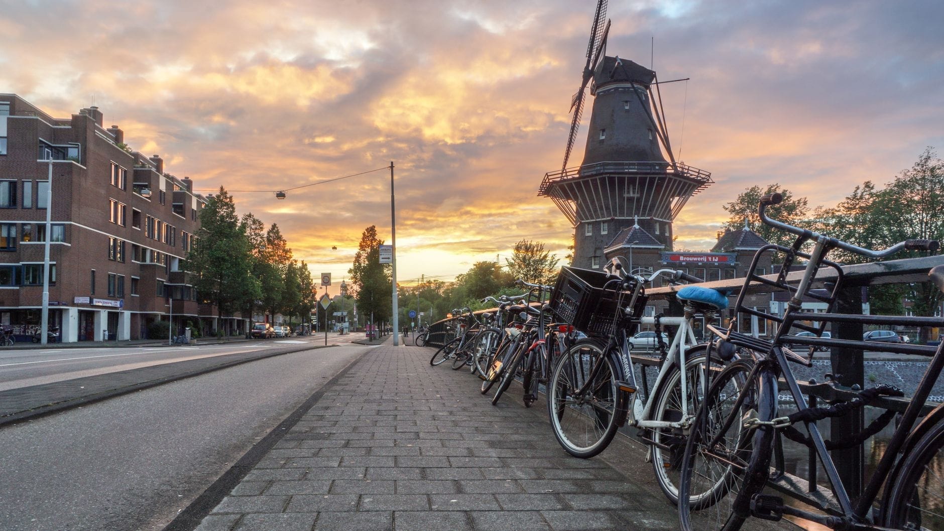 Escena callejera de Ámsterdam al atardecer con el molino De Gooyer al fondo, flanqueado por bicicletas y edificios históricos, con todo el encanto urbano holandés.