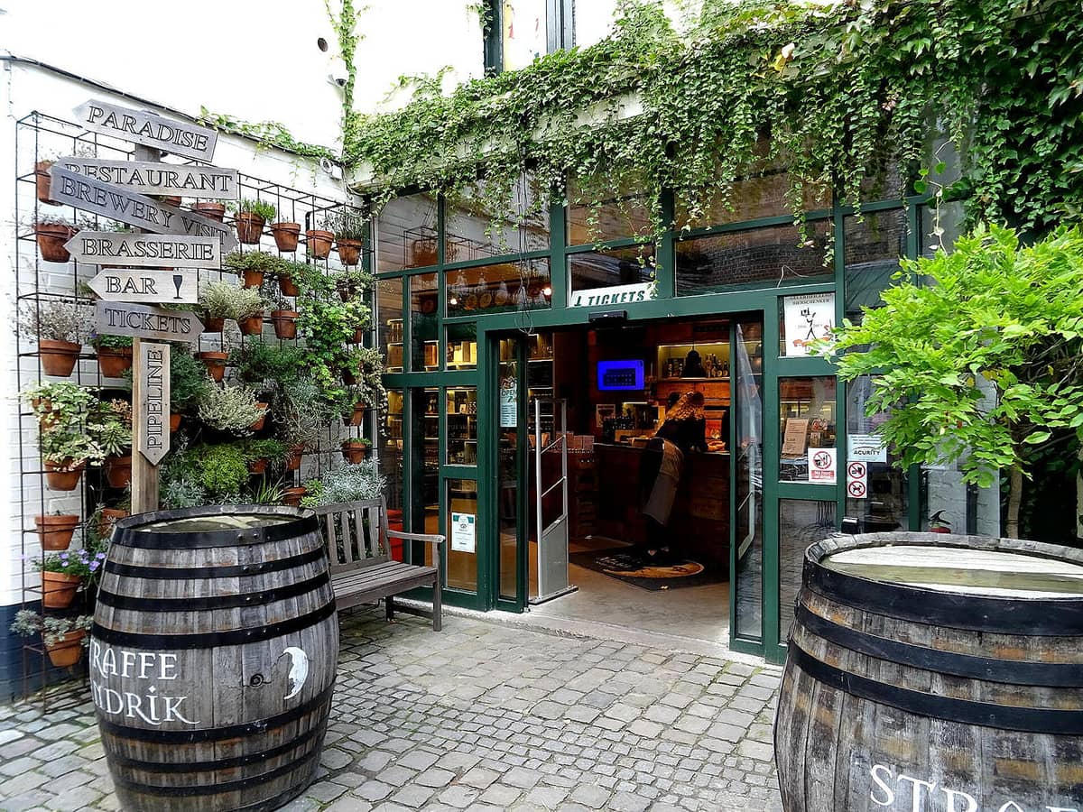 Entrance to De Halve Maan Brewery in Bruges with greenery, ticket office, and barrels labeled Straffe Hendrik on cobblestone courtyard.