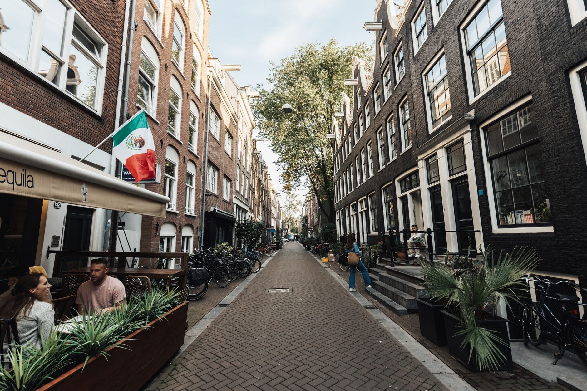 Encantadora calle de Ámsterdam en De Negen Straatjes con edificios históricos, terrazas al aire libre, bicicletas y árboles frondosos que invitan a disfrutar de un ambiente tranquilo y relajado.