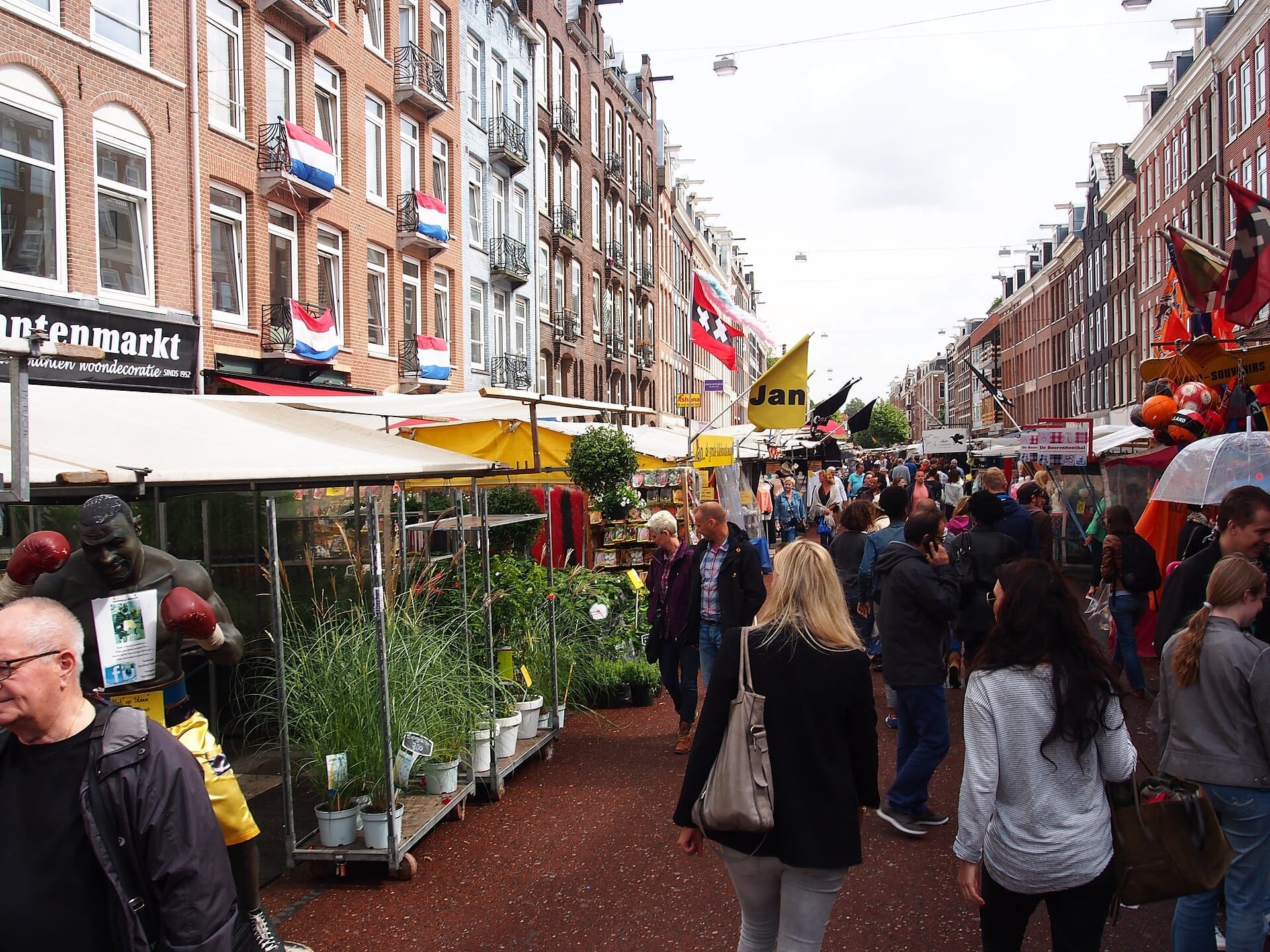 Animada escena callejera en el mercado Albert Cuyp de De Pijp, con edificios de ladrillo, banderas holandesas, coloridos puestos de mercado y multitudes de personas comprando al aire libre.