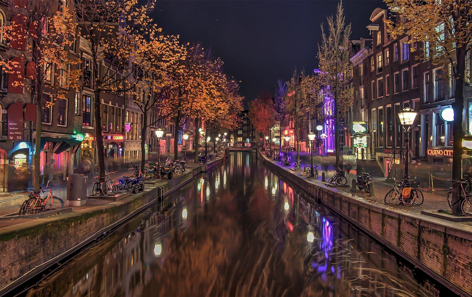 Vista nocturna del icónico canal de De Wallen en Ámsterdam, flanqueado por casas históricas, farolas encendidas, bicicletas y reflejos de colores que evocan el animado ambiente del Barrio Rojo.