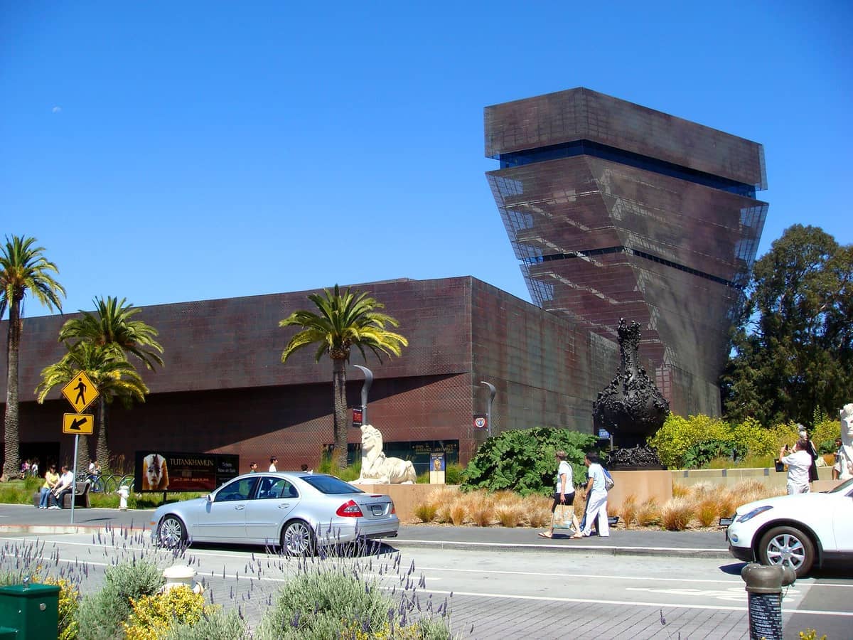 Vista exterior del de Young Museum en Golden Gate Park, con su moderna arquitectura revestida de cobre, palmeras y visitantes en un día soleado.