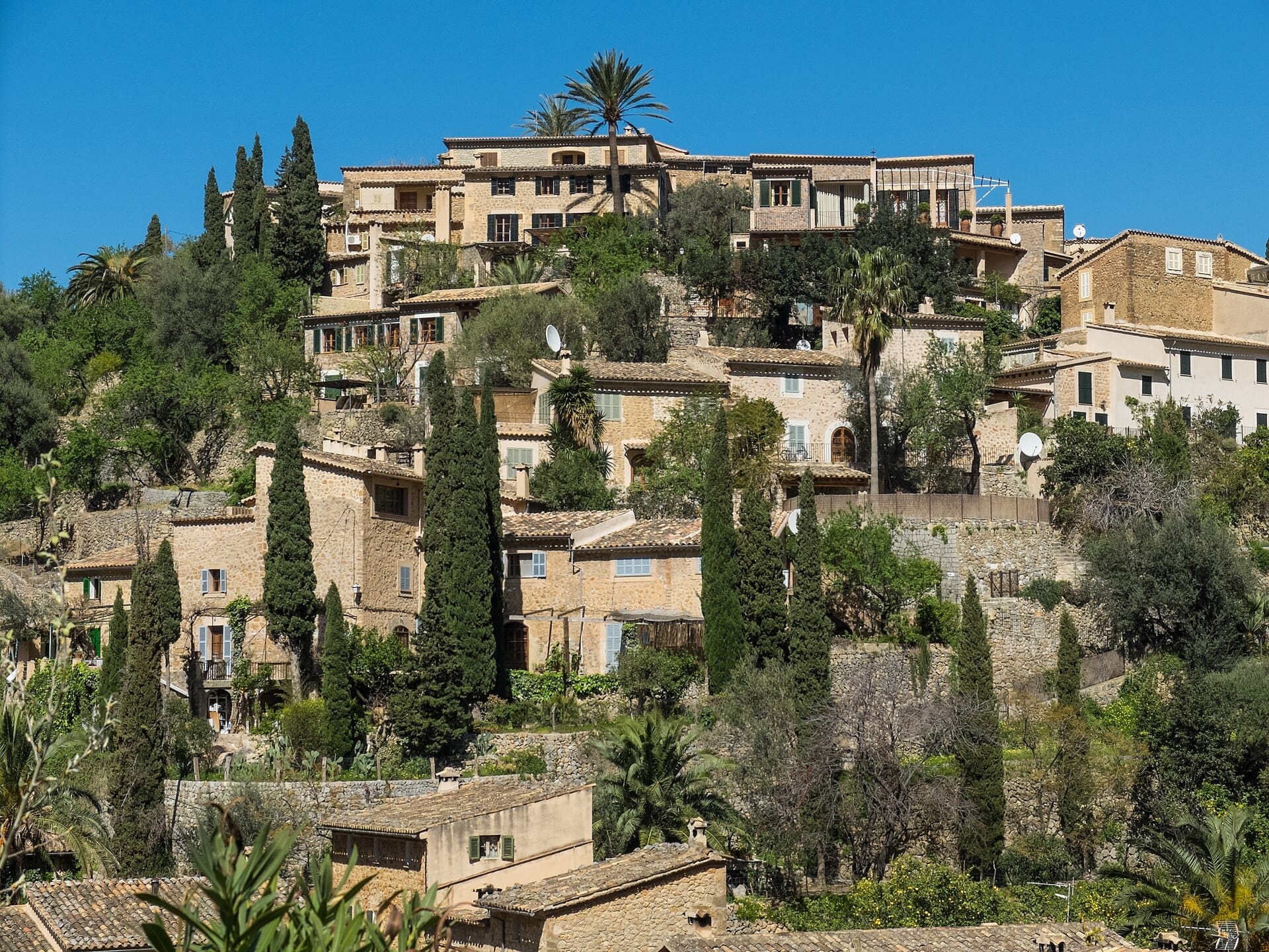 Case in pietra e cipressi salgono lungo il pendio a Deià, Maiorca, con una lussureggiante vegetazione sotto un cielo mediterraneo azzurro e sereno.