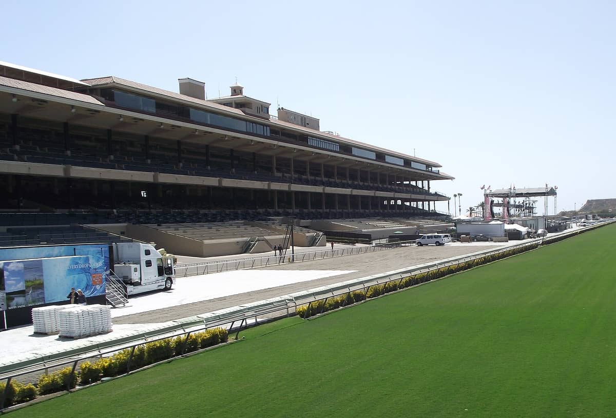 La tribuna y la pista del Hipódromo de Del Mar en California, vista desde un lateral con las gradas vacías y el prado de la pista al frente.