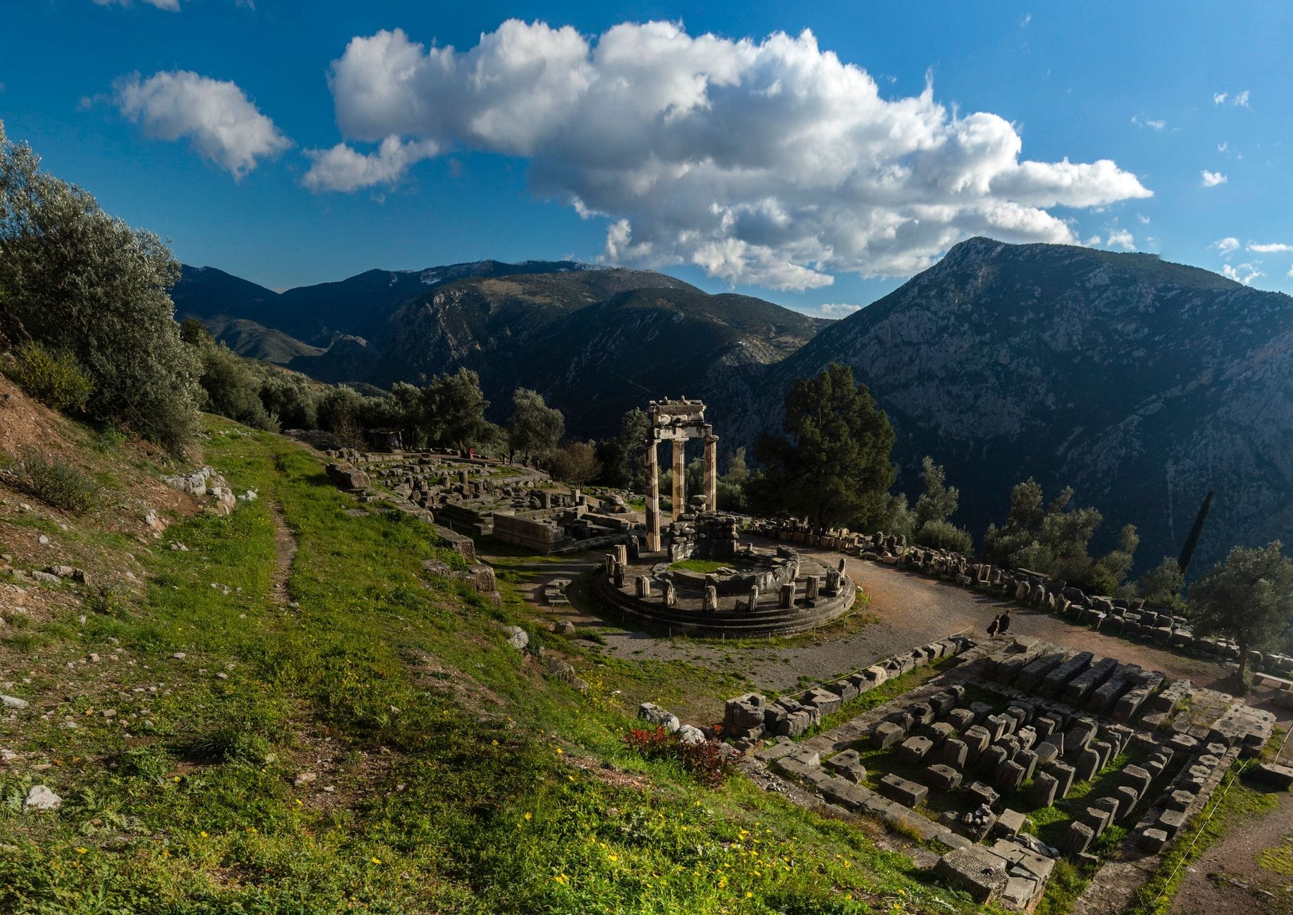 Vista panoramica del sito archeologico di Delfi con il Tholos e le rovine antiche circondati da montagne verdi sotto un cielo azzurro con nuvole.