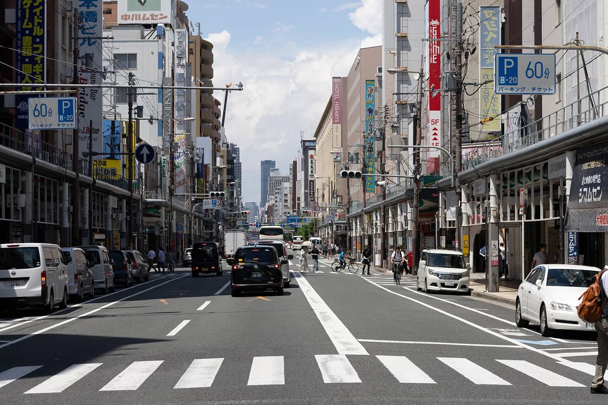 Wide street view of Nipponbashi Den Den Town in Osaka, with visible anime signs, electronics shops, and cars during daytime.