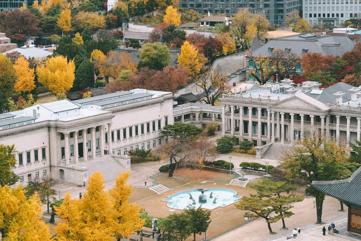 Vista aérea dos edifícios de estilo ocidental e da fonte central do Palácio Deoksugung, cercados por árvores coloridas de outono em meio aos arranha-céus modernos do centro de Seul.