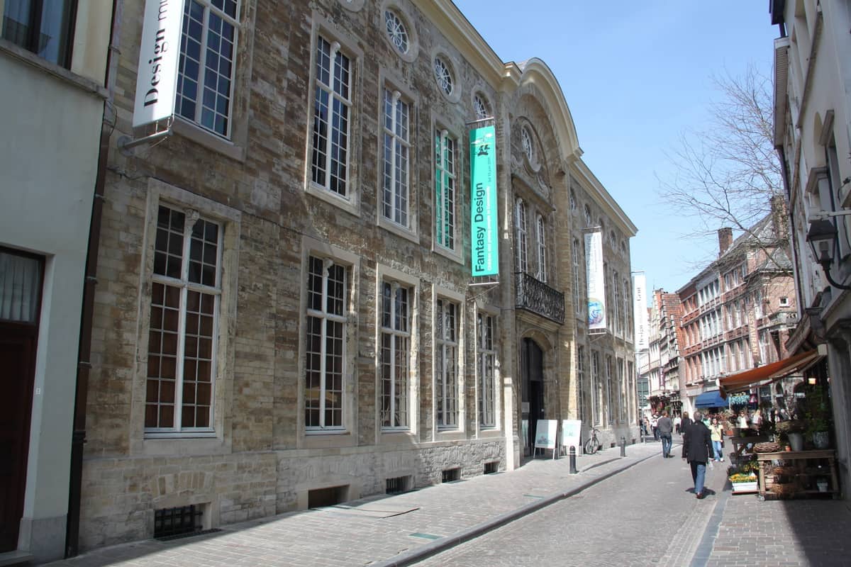 Exterior view of Design Museum Gent, a historic stone building with tall windows and banners in Ghent's old city center on a sunny day.