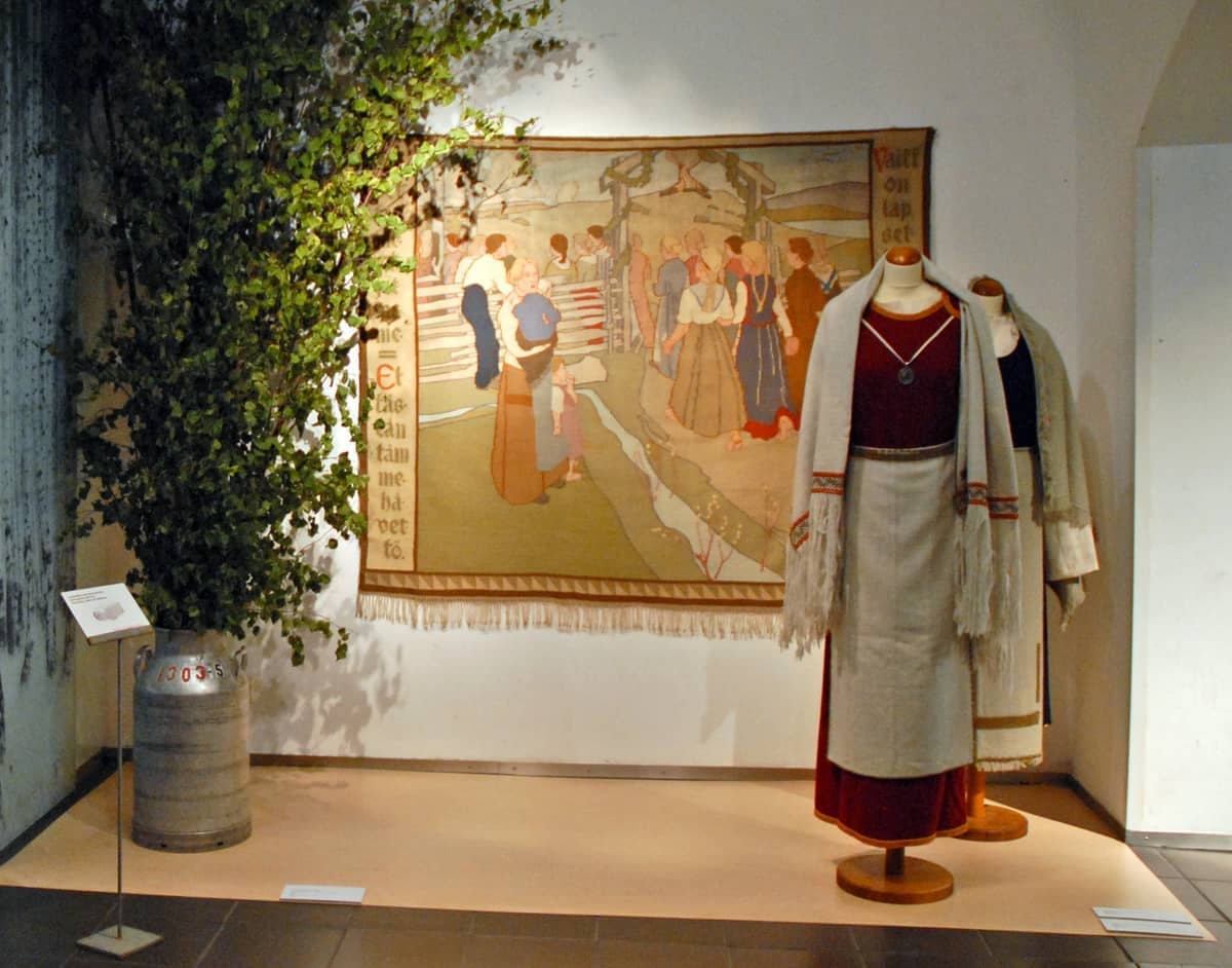 Dois manequins vestidos com roupas tradicionais finlandesas estão ao lado de uma árvore frondosa e uma grande tapeçaria no Design Museum Helsinki.