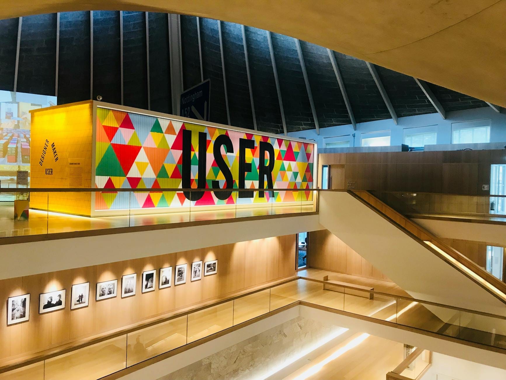 Interno del Design Museum di Londra con architettura moderna e spigolosa, illuminazione luminosa, una parete geometrica colorata e fotografie in bianco e nero esposte.