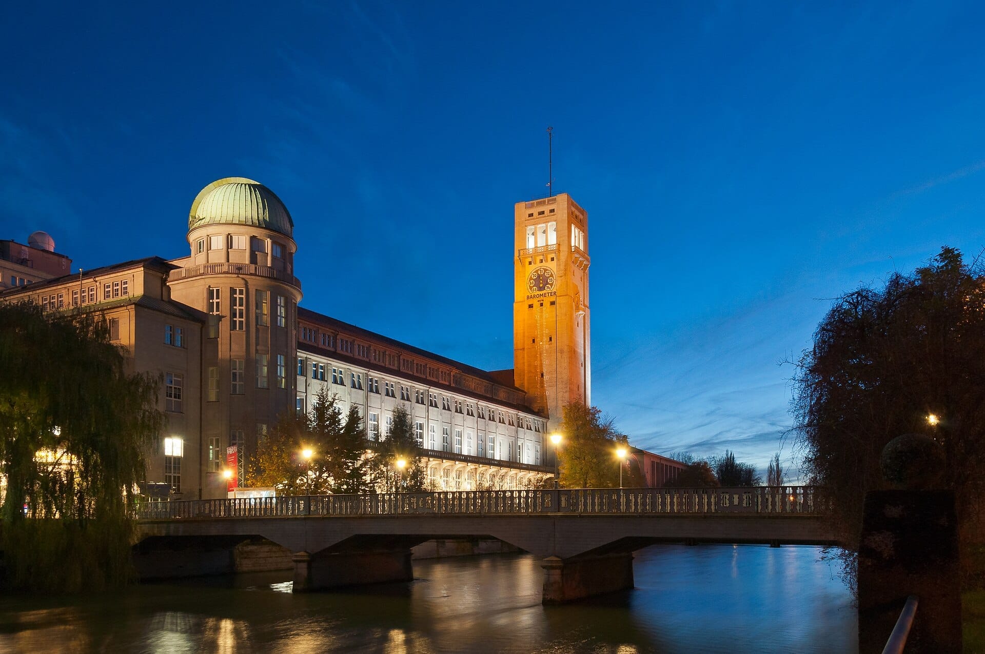 Deutsches Museum เมืองมิวนิกยามพลบค่ำ แสดงให้เห็นอาคารหลักที่มีประวัติศาสตร์ หอดูดาวทรงโดม หอนาฬิกาที่สว่างไสว และสะพานข้ามแม่น้ำ Isar
