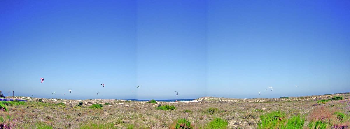 Ampie dune costiere e vegetazione mediterranea nella Devesa del Saler, con colorati kitesurfer che punteggiano il cielo azzurro limpido.