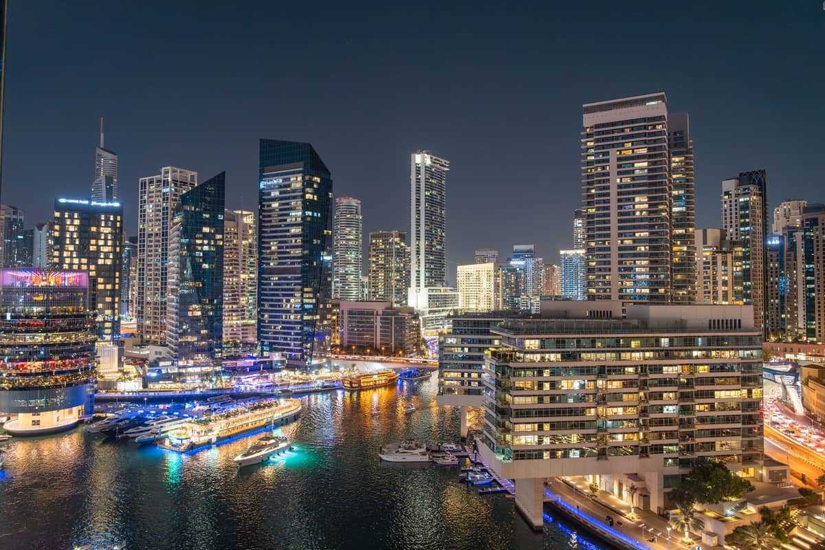 Dubai Marina bei Nacht mit beleuchteten Wolkenkratzern, glitzerndem Wasser und mehreren hell erleuchteten Booten, die entlang der Uferpromenade angelegt haben.