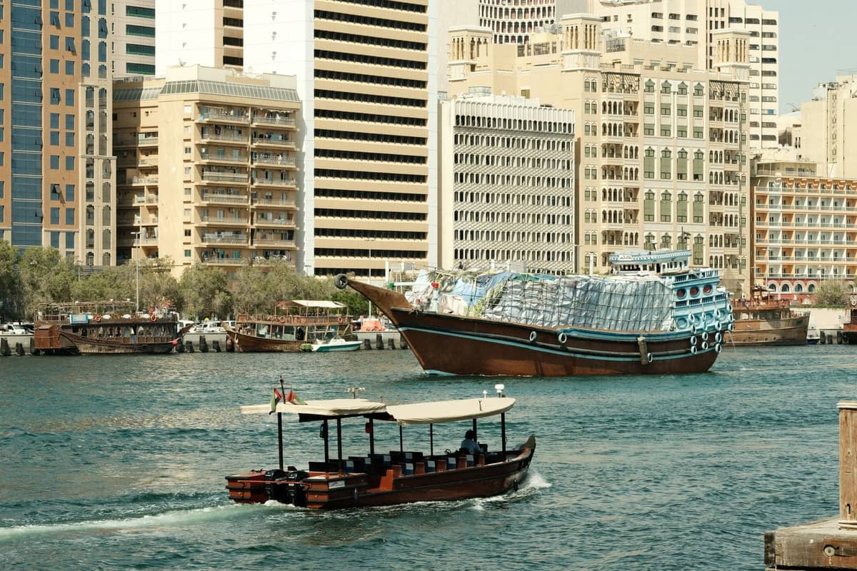 Traditionelles hölzernes Dhau-Boot auf dem Dubai Creek mit modernen Stadtgebäuden im Hintergrund bei Tageslicht.