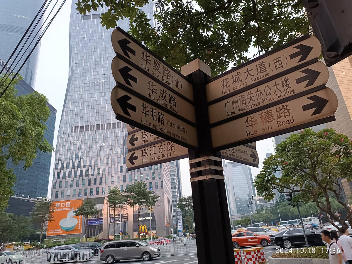 Street-level photo of Huacheng Avenue district in Guangzhou, with bilingual direction signs, high-rise modern buildings, and people walking in the city center.