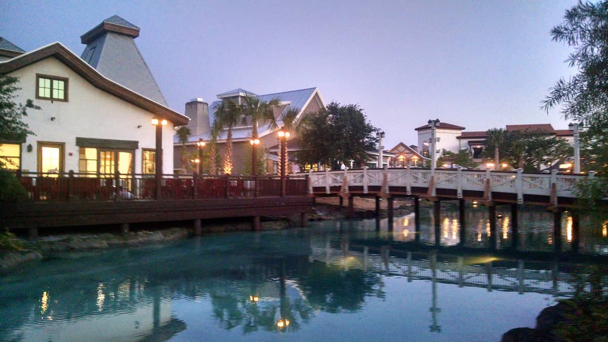 Disney Springs in der Abenddämmerung: beleuchtete Gebäude am Wasser, eine erleuchtete Promenade, Palmen und deren Spiegelungen im blauen See – die Atmosphäre des Entertainment-Viertels perfekt eingefangen.