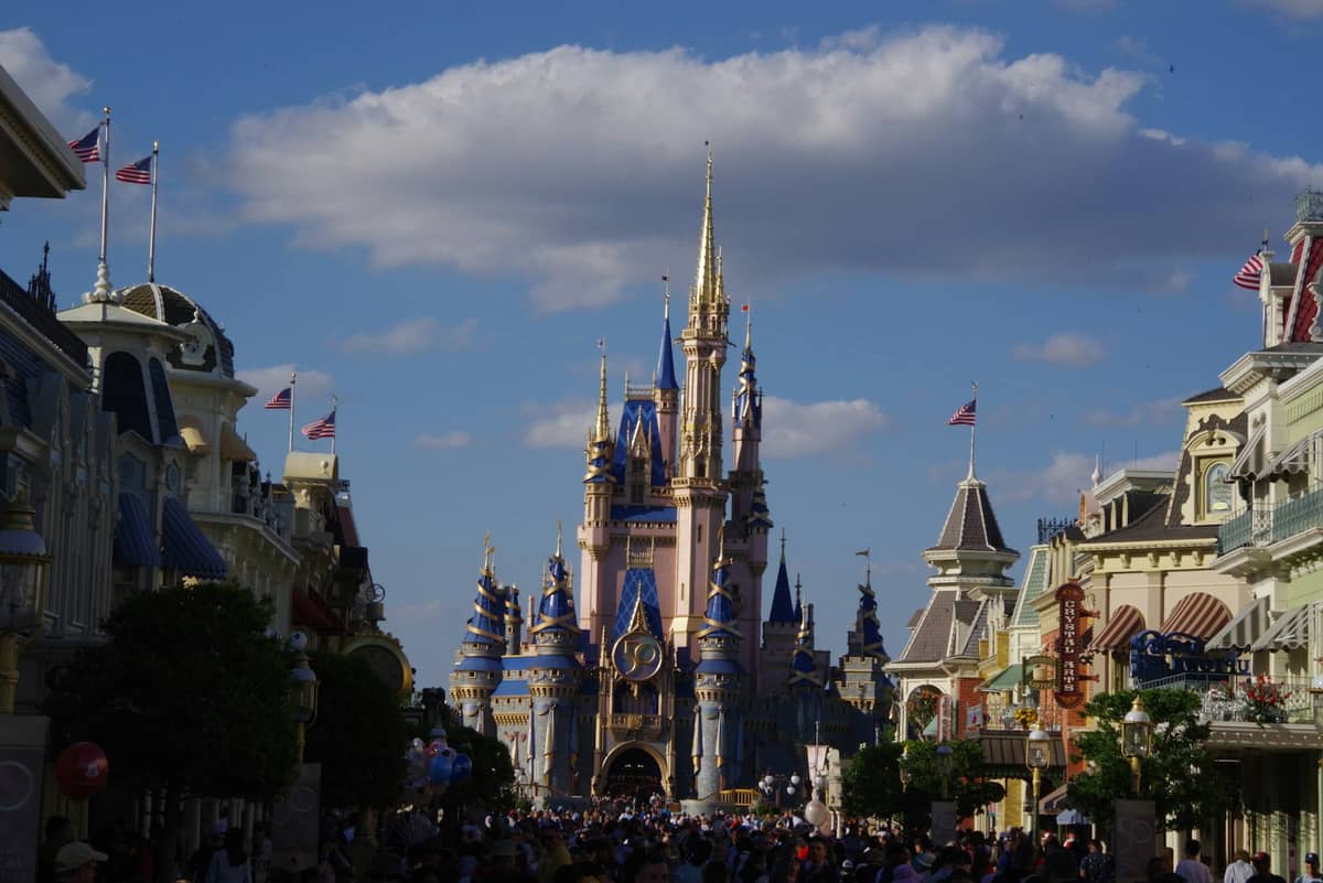 Multitudes caminando por Main Street USA hacia Cinderella Castle en Magic Kingdom bajo un cielo azul con una gran nube encima.