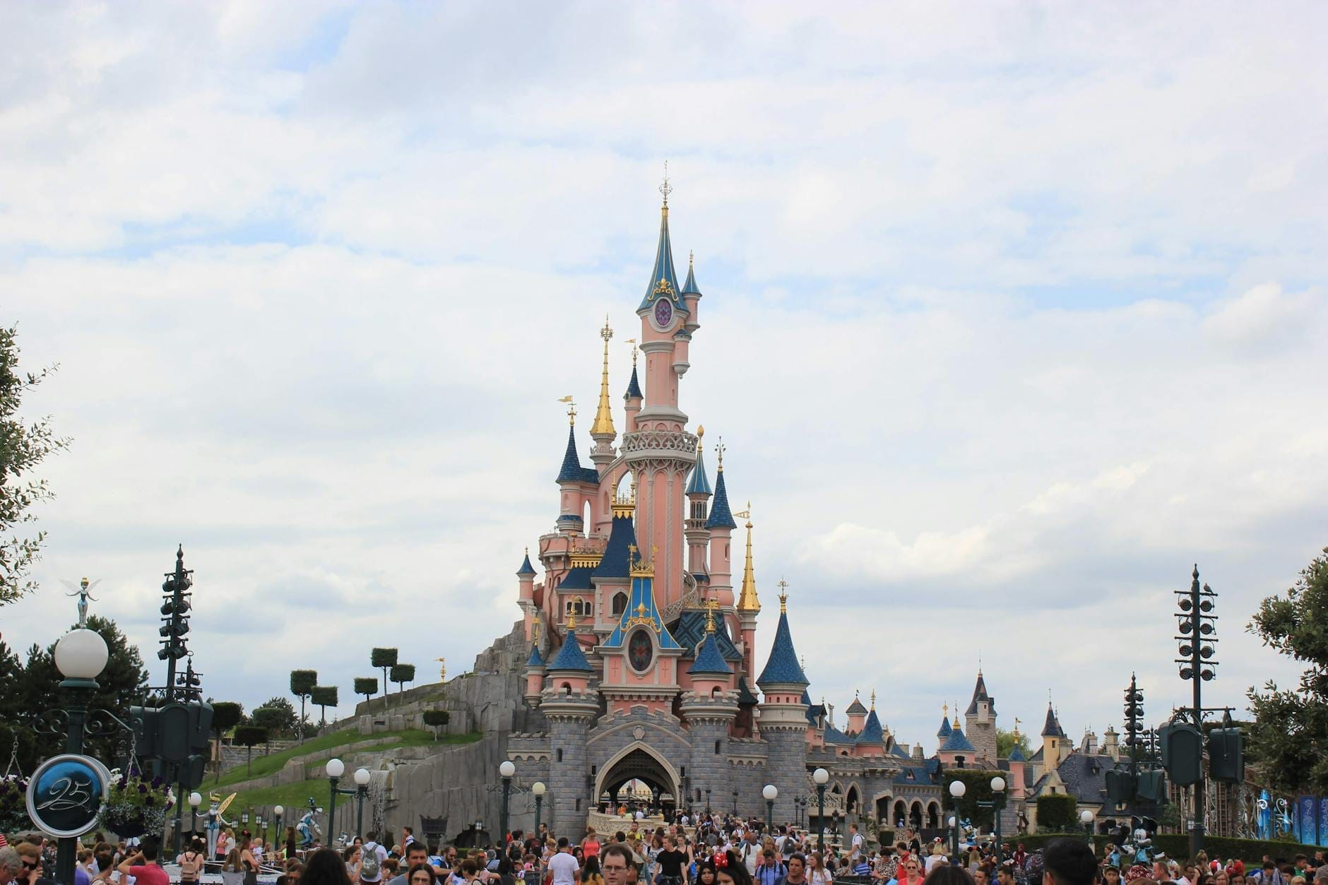 Grande folla davanti all'iconico Castello della Bella Addormentata rosa a Disneyland Paris, sotto un cielo luminoso con nuvole sparse, tra entusiasmo e meraviglia.