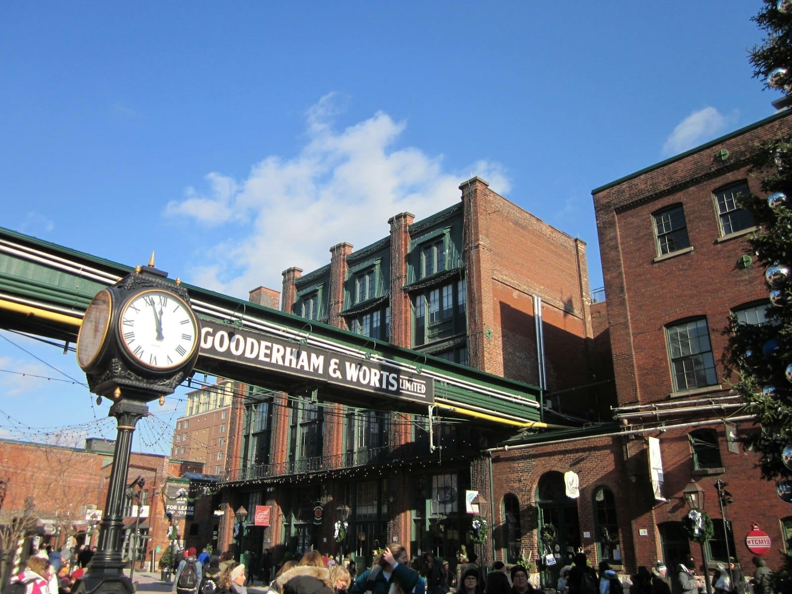 บรรยากาศคึกคักใน Distillery Historic District มีอาคารอุตสาหกรรมอิฐแดงยุควิคตอเรีย ป้าย Gooderham & Worts และนาฬิกาประดับใต้ท้องฟ้าแจ่มใส