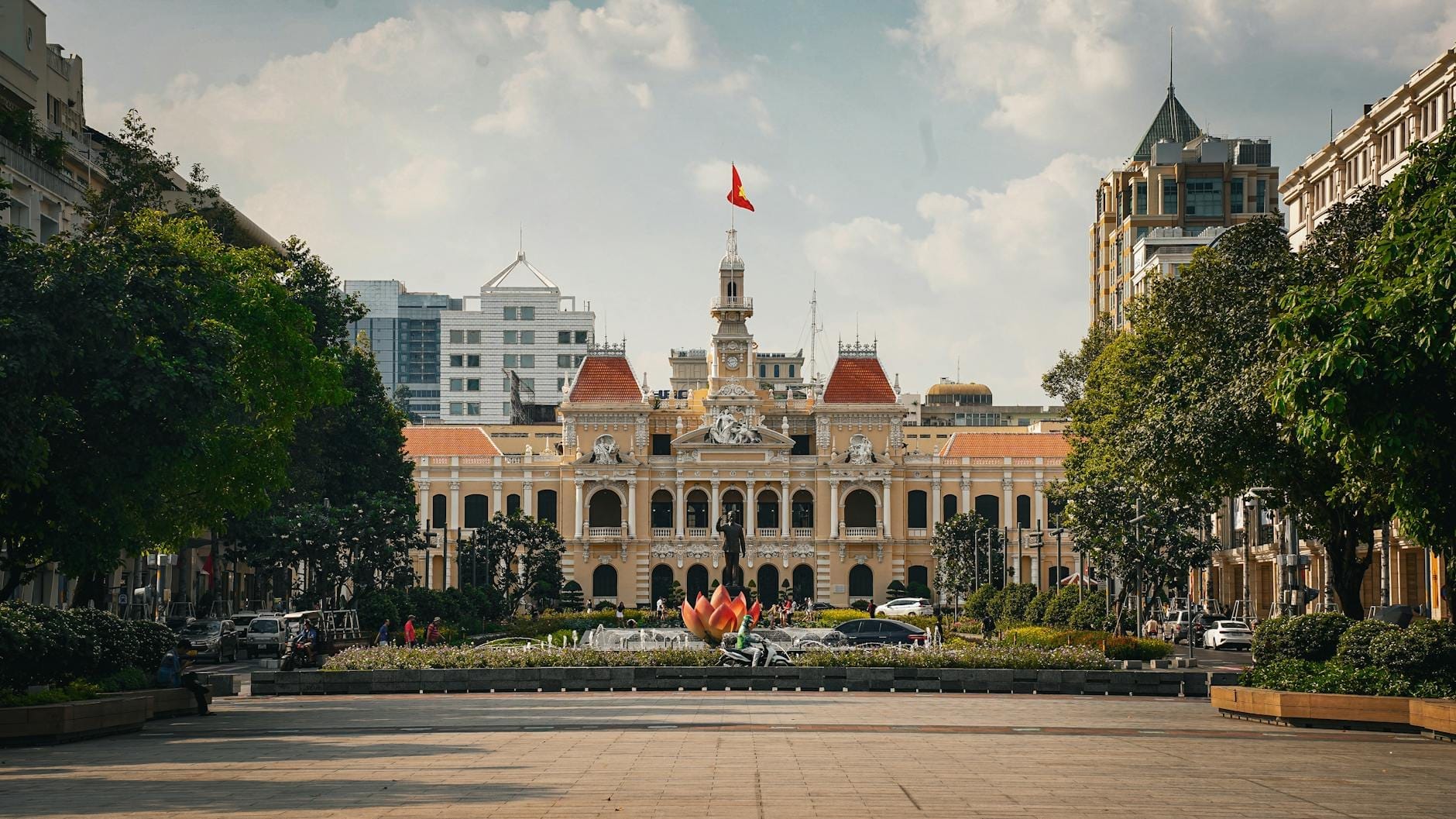 Vista panorámica del Ayuntamiento de Ho Chi Minh con un cielo azul intenso, rodeado de árboles, una plaza con fuente de loto y edificios modernos al fondo.