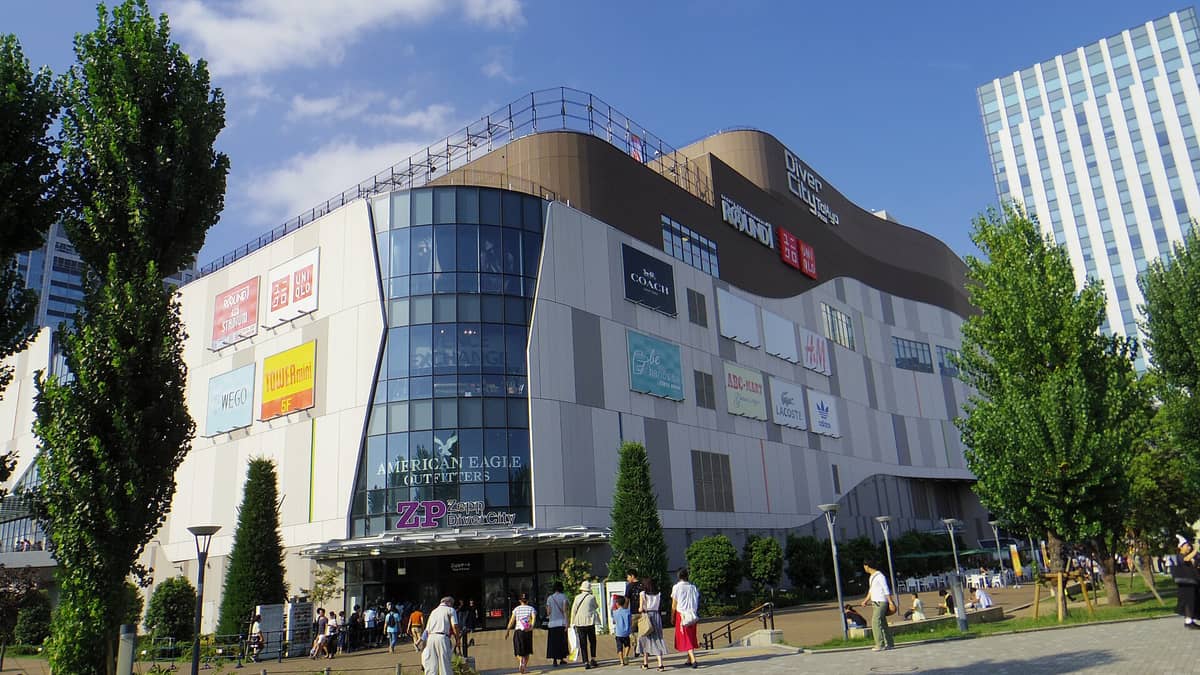 Exterior view of DiverCity Tokyo Plaza with its recognizable modern facade, large brand signs, and people walking in the outdoor plaza under a clear blue sky.
