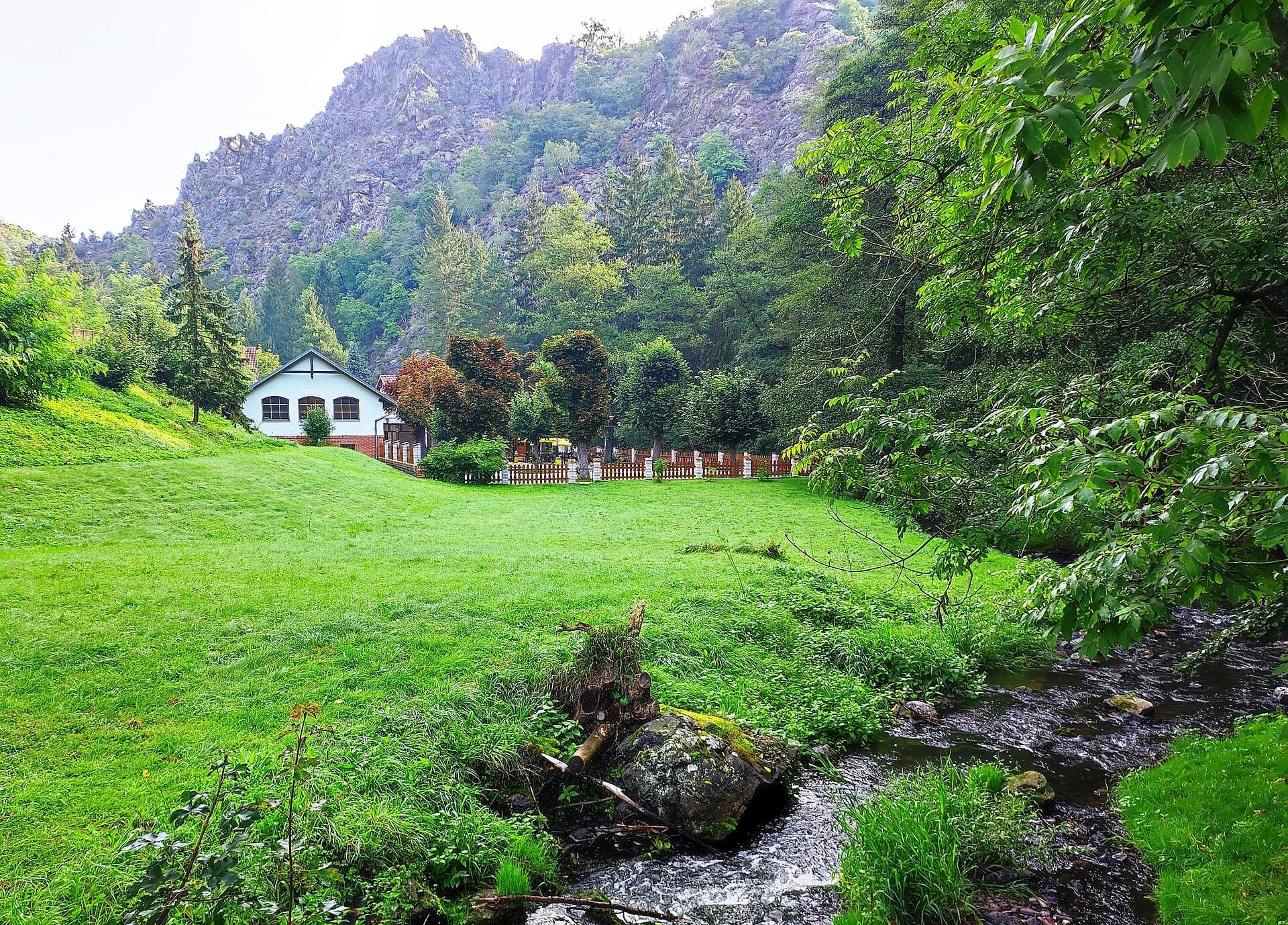 Ein kleines weißes Häuschen steht auf einer üppig grünen Wiese im Naturschutzgebiet Divoká Šárka, umgeben von felsigen Klippen und dichtem Baumbestand.