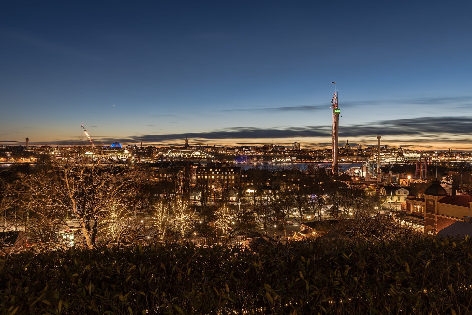 วิว Djurgården ยามพลบค่ำ เห็นสวนสนุก Gröna Lund ไฟสว่าง ริมน้ำ และฉากหลังของเมืองสตอกโฮล์ม
