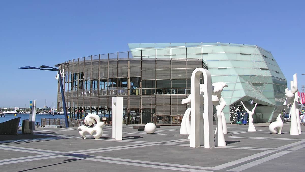 Plaza frente al agua en Docklands, Melbourne, con llamativas esculturas públicas blancas frente a un edificio de vidrio y acero con vistas al Victoria Harbour.