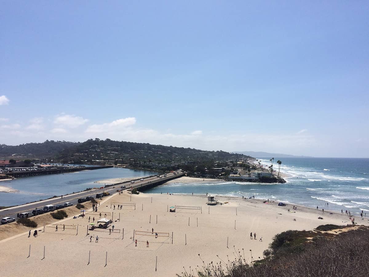 มุมมองกว้างของ Dog Beach Ocean Beach แสดงให้เห็นสนามวอลเลย์บอลทราย แม่น้ำ San Diego เนินเขาในระยะไกล และคลื่นใต้ท้องฟ้าสีฟ้าแจ่มใส