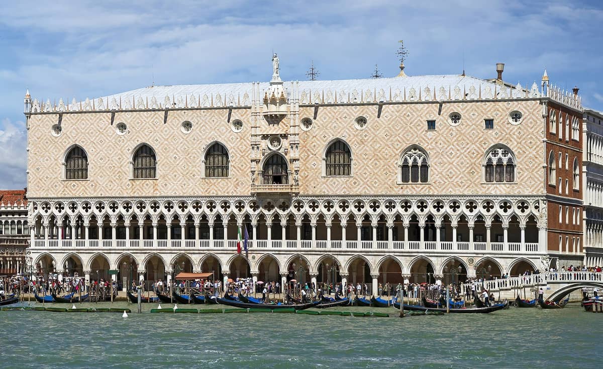 Weitwinkelaufnahme des Dogenpalastes (Palazzo Ducale) in Venedig vom Wasser aus, mit seiner aufwendig verzierten gotischen Fassade, den Bogenfenstern und einer Menschenmenge am Kanal.