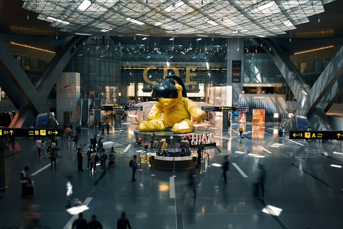 Belebtes Inneres des Hamad International Airport, Doha, mit der ikonischen riesigen gelben Teddybär-Skulptur und Reisenden, die durch die Halle laufen.