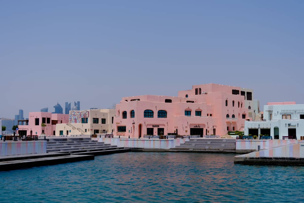 Bunte Pastellgebäude am Wasser im Katara-Viertel in Doha, mit dem Golfwasser und modernen Wolkenkratzern im Hintergrund.
