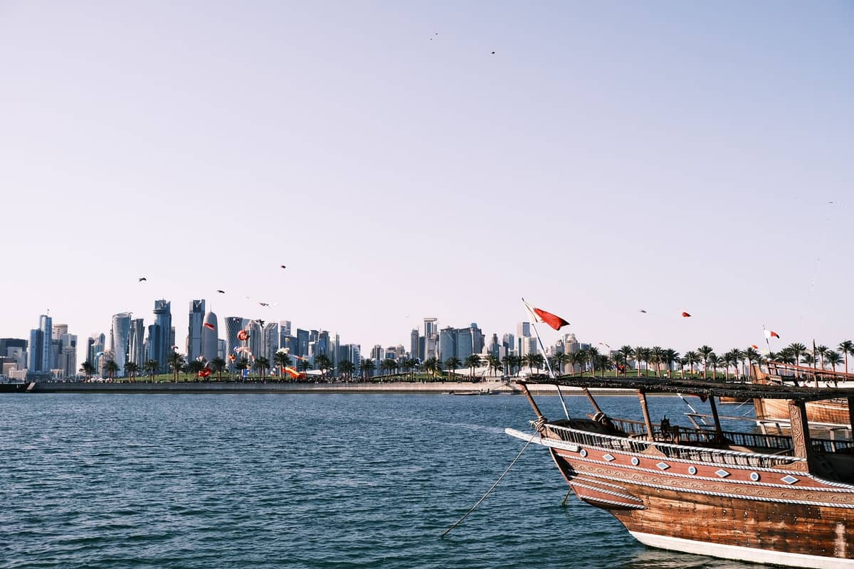 Traditionelles hölzernes Dhau-Boot, das in der Bucht von Doha vertäut ist, mit der modernen Skyline von West Bay im Hintergrund unter klarem Tageshimmel.