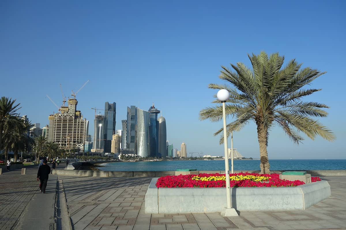 Die Uferpromenade der Doha Corniche mit Palmen, Blumenbeeten und Spaziergängern am Wasser, im Hintergrund die moderne West-Bay-Skyline.