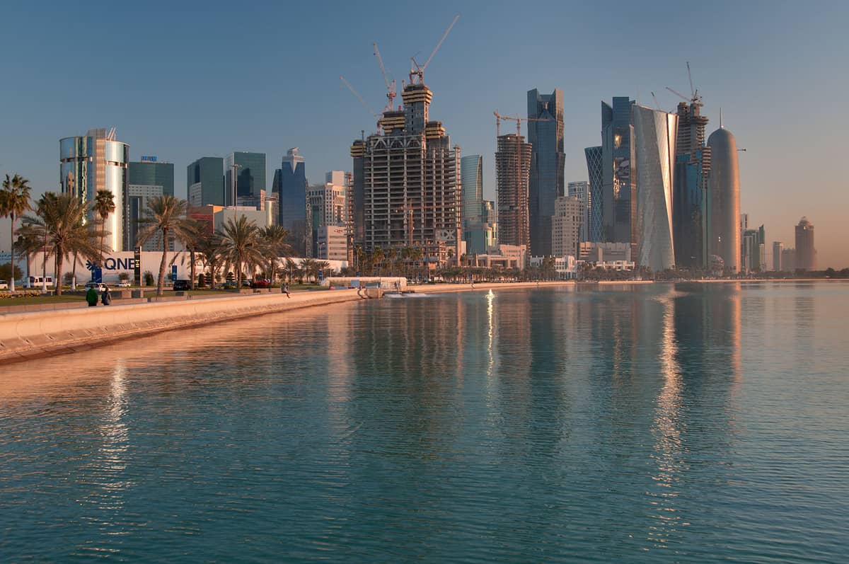 Weitwinkelansicht der Doha Corniche bei Sonnenuntergang mit Palmen, die modernen West Bay-Wolkenkratzer spiegeln sich im ruhigen Buchtenwasser.