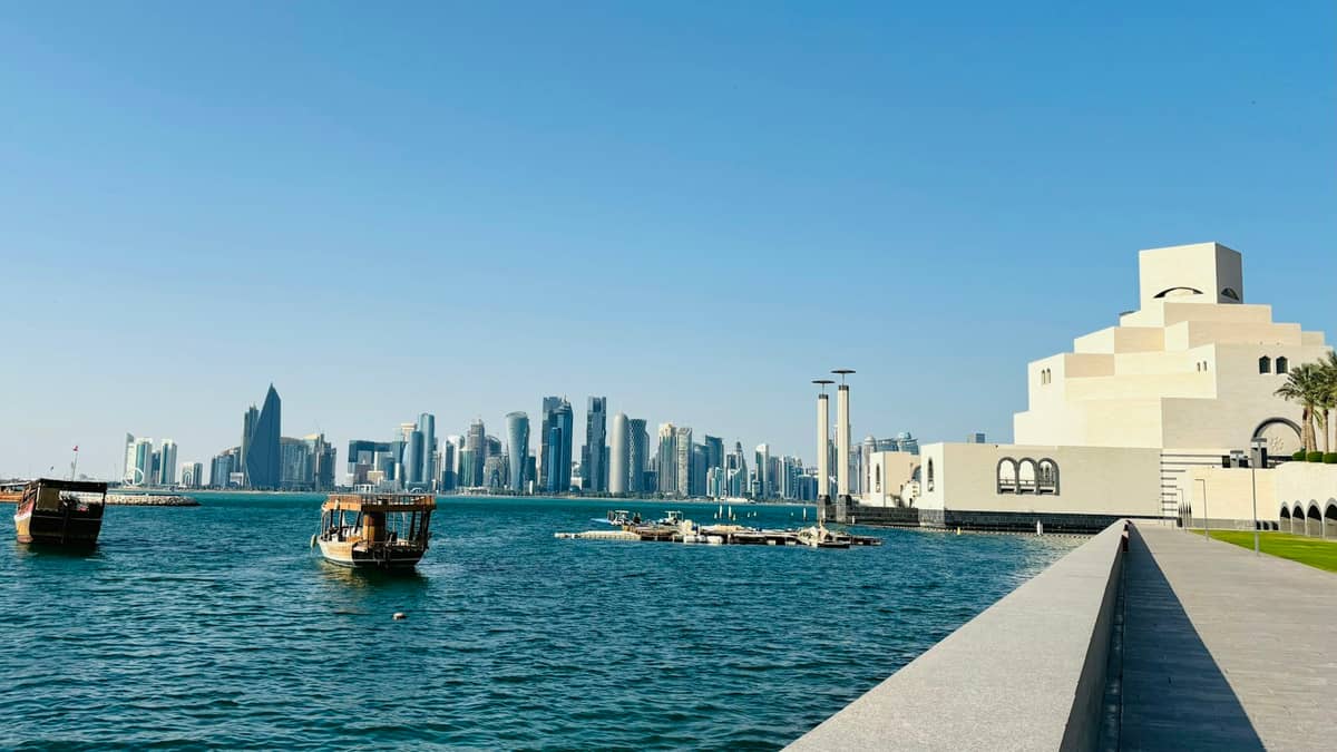 Skyline von Doha jenseits der Bucht mit Dau-Booten und dem Museum für Islamische Kunst neben der breiten Corniche-Promenade an einem sonnigen Tag.