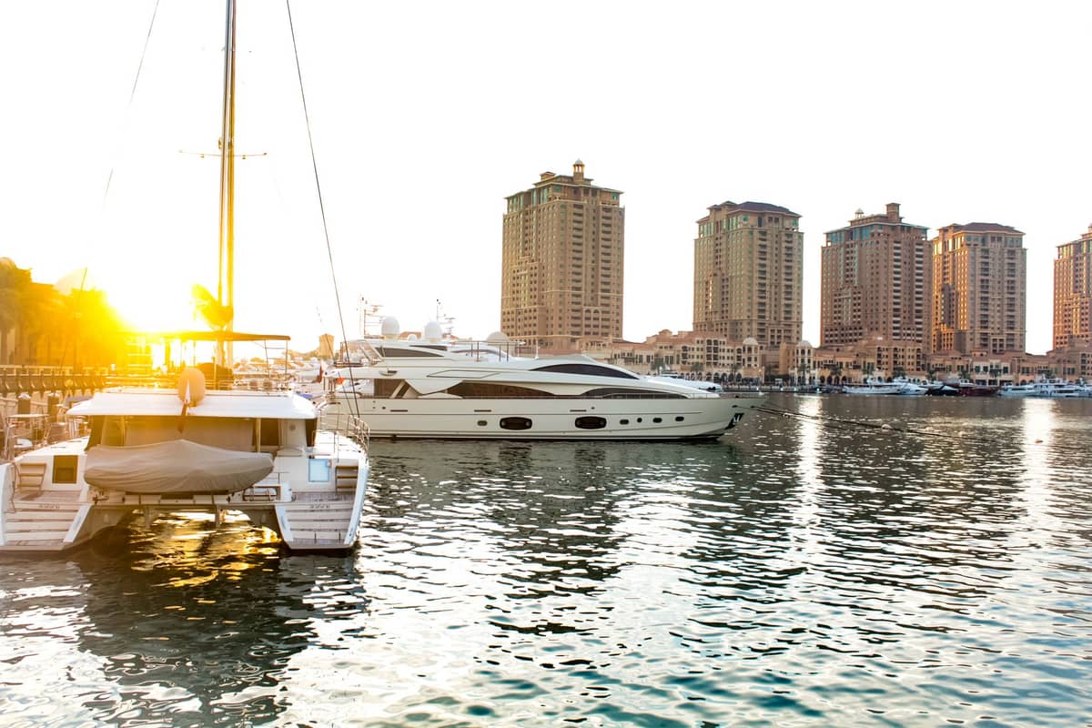 Luxusyachten und Hochhausresidenzen an der Marina von The Pearl-Qatar in Doha bei Sonnenuntergang, mit klarem Wasser, das das Sonnenlicht widerspiegelt.