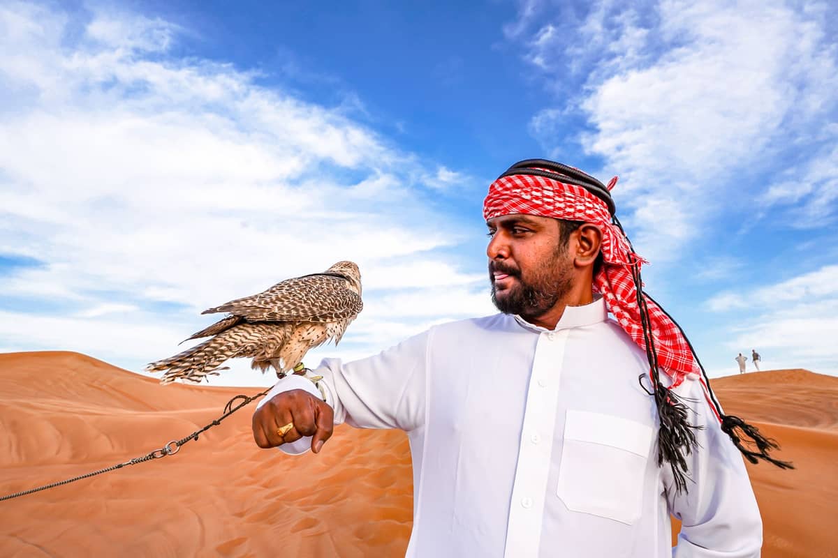 Mann in weißem Thobe und rotem Keffiyeh hält einen Falken auf seinem Arm in der Wüste unter blauem Himmel.