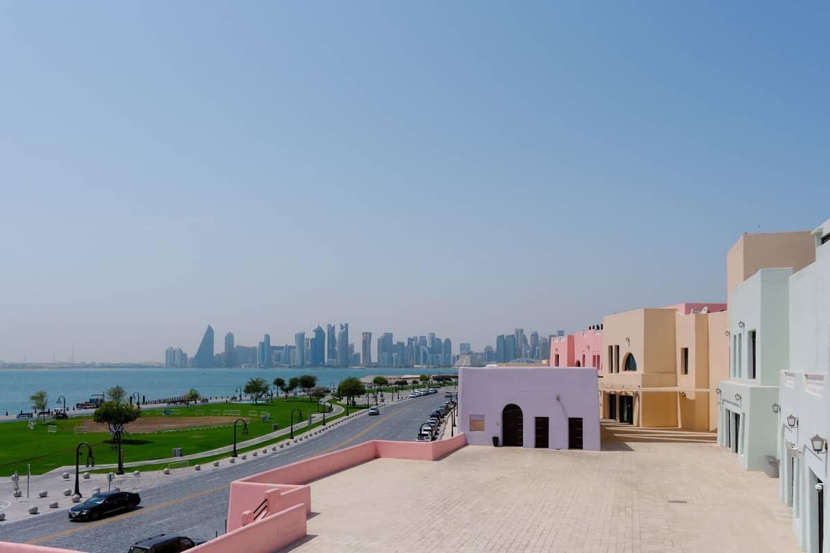 Weiter Blick bei Tag auf die Doha Corniche, bunte Katara-Gebäude rechts, Uferpark und die markante Skyline von West Bay in der Ferne unter klarem Himmel.