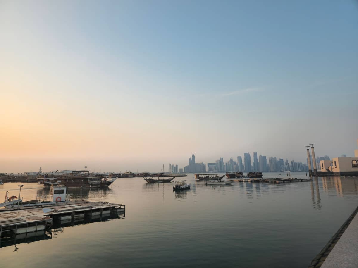 Morgenansicht des Doha Fischmarkts und Old Doha Ports mit vertäuten Booten, ruhigem Wasser und der Skyline der Stadt im dunstigen Hintergrund.