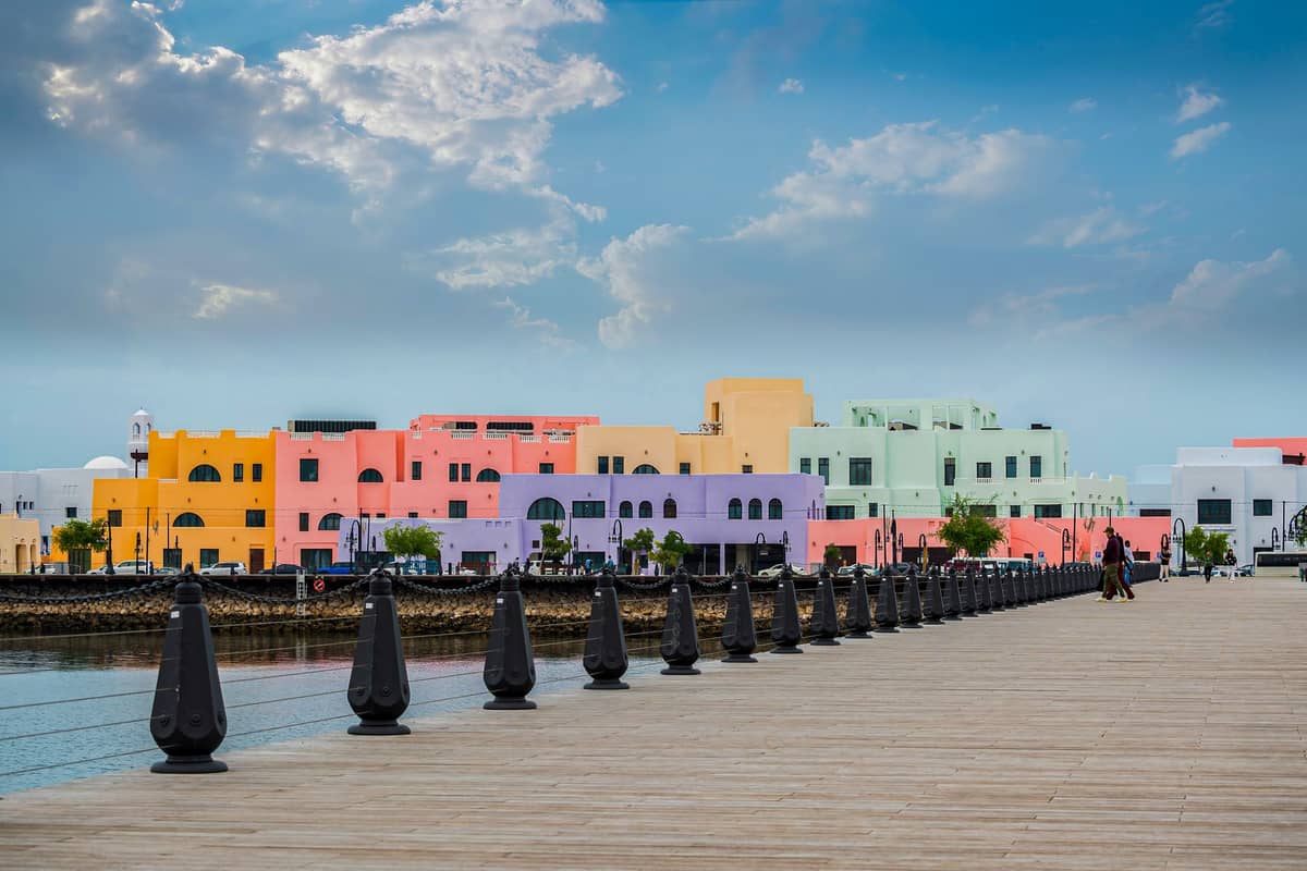 Breite Promenade mit bunten Pastellgebäuden am Alten Hafen von Doha, heller Himmel und Wasser im Vordergrund, ein wichtiges Stadterneuerungsgebiet in der Nähe des Fischmarkts.