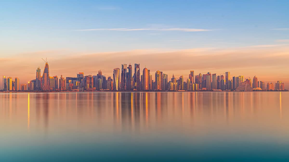 Weiter Blick auf Dohas Skyline über ruhiges Wasser bei Sonnenuntergang, mit pastellfarbenen Reflexionen auf der glatten Oberfläche und klarem Himmel.