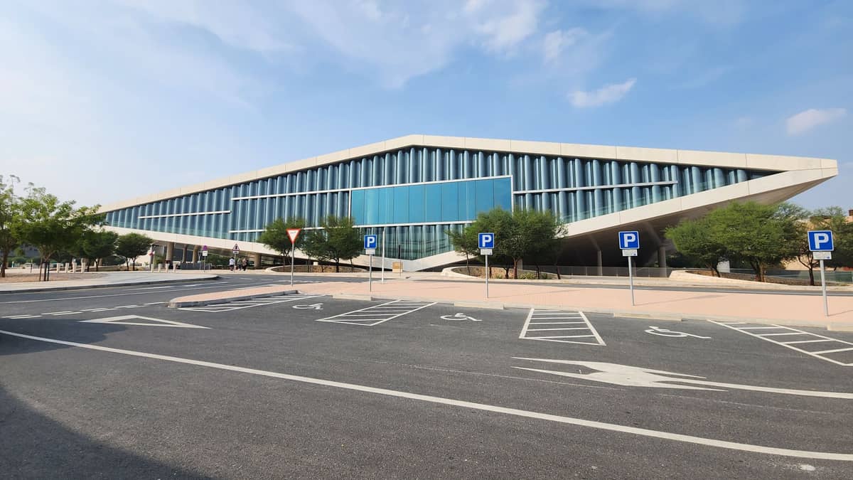 Breite Horizontalansicht des Gebäudes der Qatar National Library in Doha, mit moderner Glasfassade und leerem Parkplatz im Vordergrund.