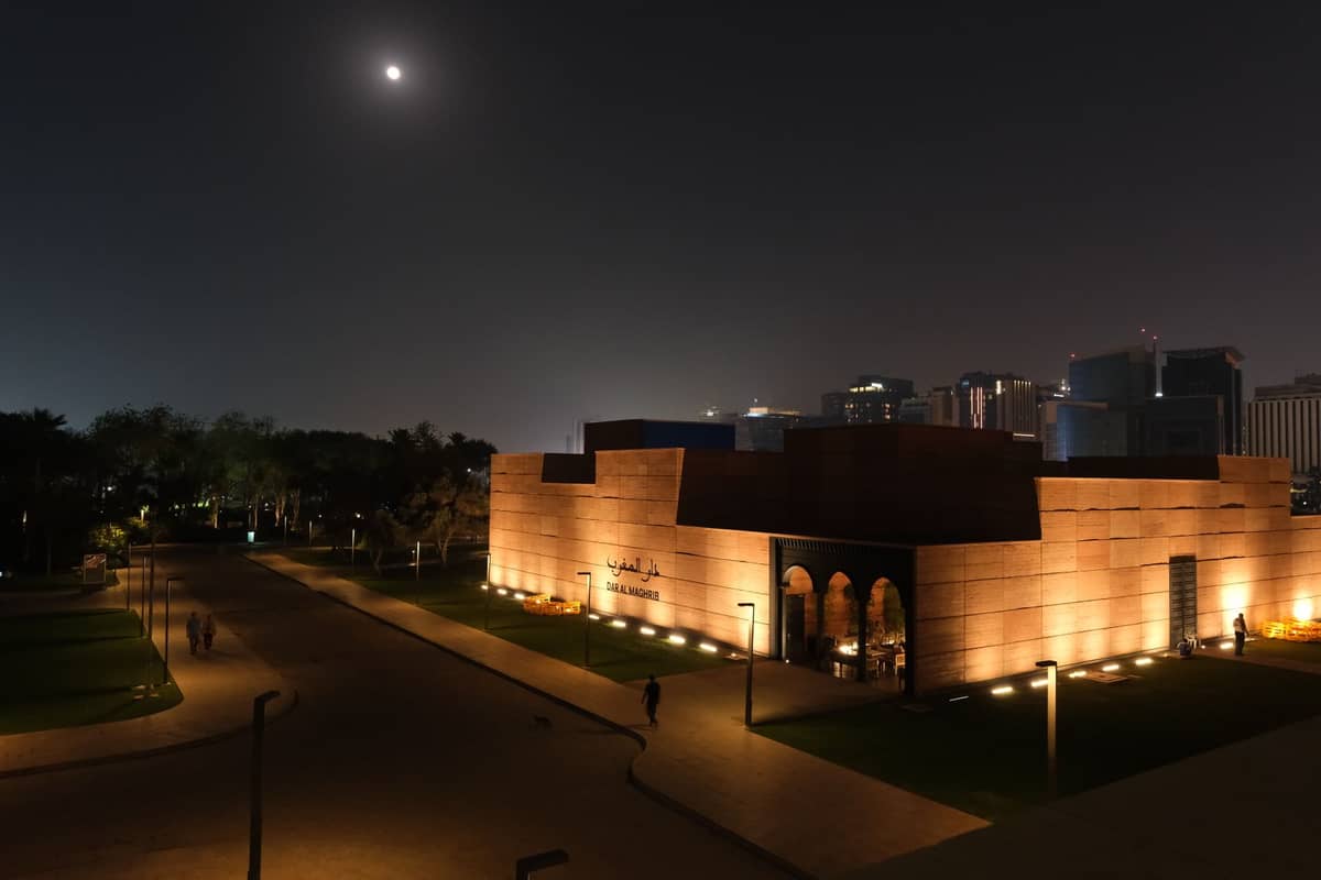 Nächtliche Straßenansicht in Doha mit einem beleuchteten modernen Gebäude, wenigen Fußgängern und der Skyline der Stadt im Hintergrund unter dem Mond.