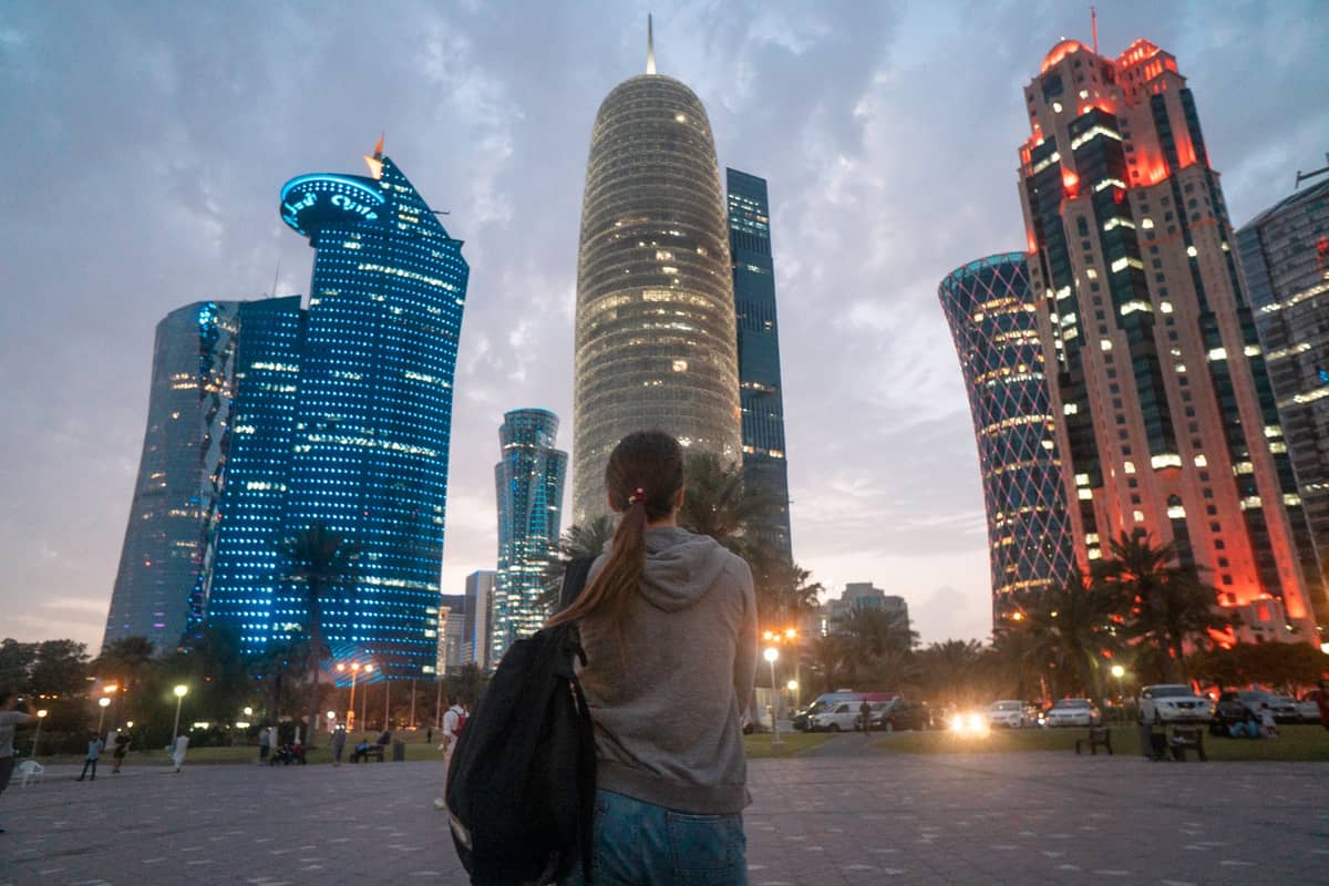 Person steht und blickt auf die beleuchteten Türme von West Bay in Doha in der Dämmerung, mit bunten Lichtern an modernen Wolkenkratzern.