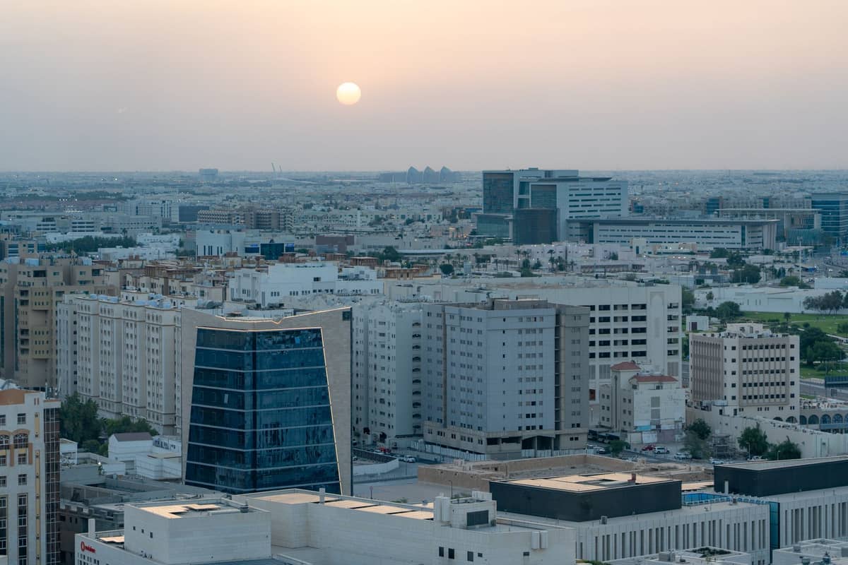 Weitwinkelansicht der Skyline von Doha bei Sonnenuntergang mit tief stehender Sonne in dunstigem Himmel über modernen Gebäuden.