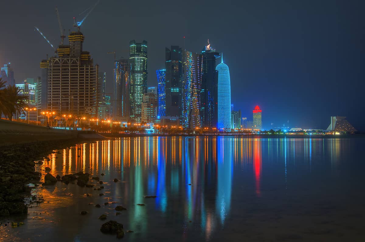 Nachtansicht der Doha Corniche Uferpromenade mit Palmen, beleuchterter Skyline und farbenfrohen Spiegelungen in der Bucht.