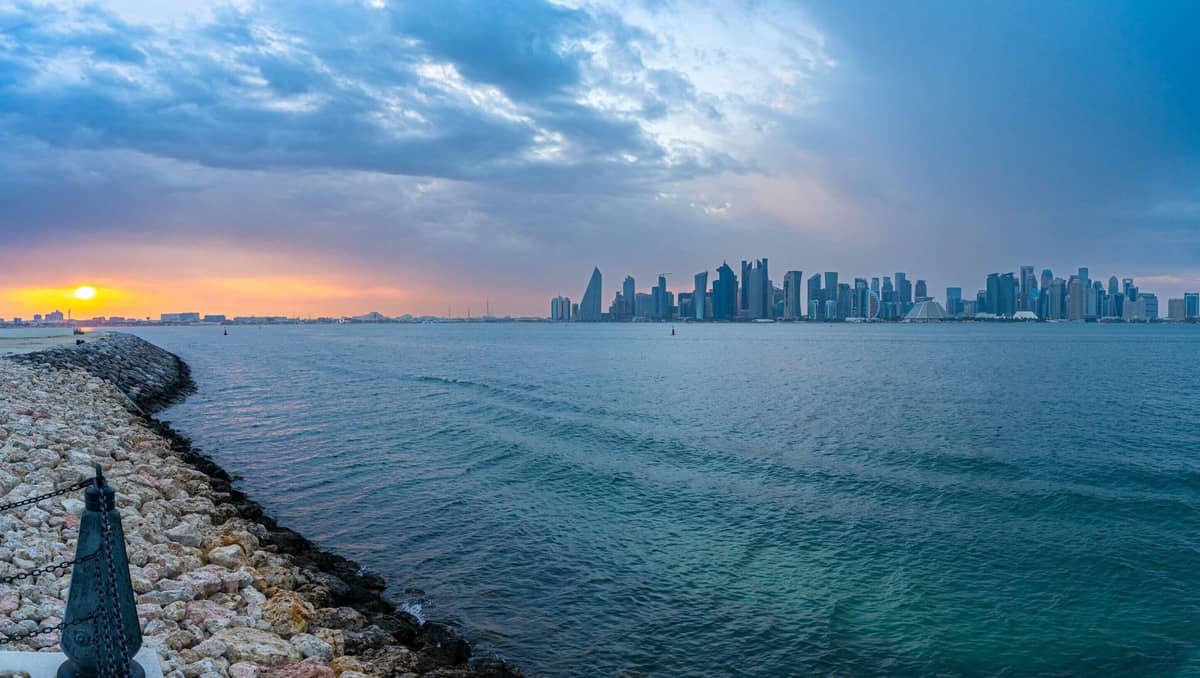 Panoramablick auf die ikonische Skyline von Doha mit modernen Türmen jenseits der Bucht bei Sonnenuntergang, gesehen von einer felsigen Promenade entlang der Corniche-Uferpromenade.
