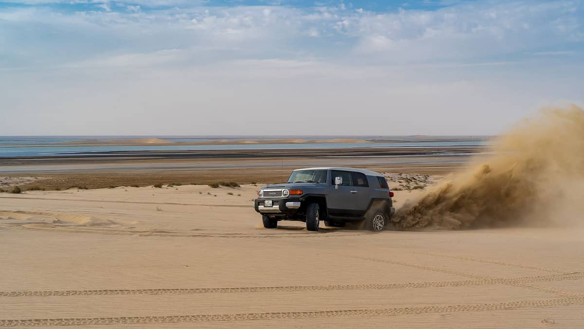 SUV fährt schnell über Sanddünen mit beeindruckenden Reifenspuren, Staubwolke dahinter und flachem Binnenmeer unter strahlend blauem Himmel